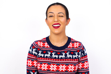 Sticker - Young beautiful Arab woman wearing Christmas sweater against white wall blinking eyes with pleasure having happy expression. Facial expressions and people emotions concept.