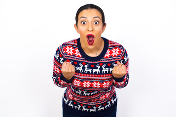 Wall Mural - Joyful excited lucky Young beautiful Arab woman wearing Christmas sweater against white wall, cheering, celebrating success, screaming yes with clenched fists