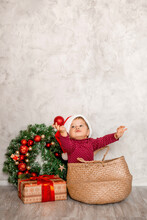 Sweet Baby Santa Sits In A Wicker Basket With A Gift Box And A Christmas Wall On A Gray Background. Christmas Concept, Text Space