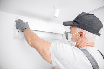 Technician man in medical mask worker installs and clear air conditioner on white wall