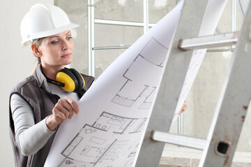 woman construction worker builder looking at bluprint, wearing helmet and hearing protection headphones in building site indoors background