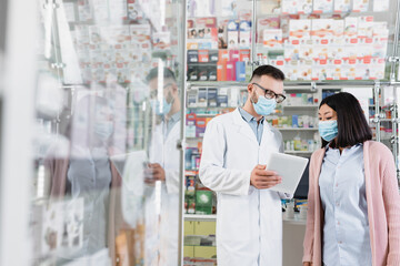 Wall Mural - pharmacist in medical mask holding digital tablet near asian woman in drugstore