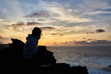 Wall Mural - Hispanic female sitting on a rocky seashore and observing the beautiful view of Gran Canaria island