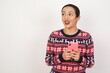 © Roquillo - Portrait of  Young beautiful Arab woman wearing Christmas sweater against white wall,  holding in hands showing new cell