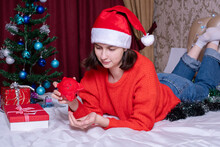 Teen Girl In A Christmas Hat And A Red Sweater Pulls Out Coins From A Red Piggy Bank To Shop For Christmas Gifts