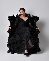 Canvas Print - full length portrait of  woman wearing black gothic dress, sitting on a ornate black armchair. Seated pose, against a studio background.