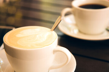 Black coffee in a white ceramic cup and a cup of cappuccino with foam on a wooden table in the morning sun. Selective focus. Breakfast in a cafe