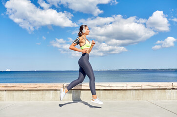 fitness, sport and healthy lifestyle concept - young woman with headphones and smartphone in armband running at seaside