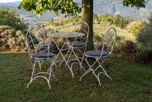 Set Of Four Outdoor Ornate Chairs And Table Next To A Tree With A View Across An Italian Valley In Tuscany.