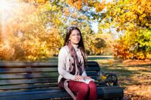 Portrait Of A Beautiful, Brunette Woman Sitting On A Bench In A Park During Autumn Time With Colorful Trees And Sunshine