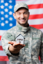 House Model In Hand Of Happy Military Man In Uniform Near American Flag On Blurred Background