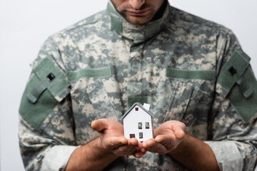 Wall Mural - house model in hands of patriotic military man in uniform on blurred background isolated on white