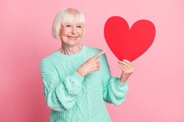 Photo portrait of adorable grandmother pointing at postcard red heart symbol valentines day isolated on pastel pink color background