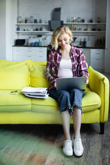 Wall Mural - smiling blonde woman in checkered shirt working from home with laptop on knees