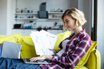 Wall Mural - blonde woman in checkered shirt typing on laptop and holding documents