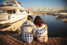 Back View Of Young Couple Sitting Together On Wooden Pier In The Port With Small Yachts Near To The Sea Water.