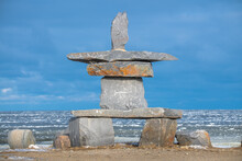 A Large Inukshuk That Sits On The Shoreline Of Hudson Bay In Churchill, Manitoba In Northern Canada. Icy Sea Ice In The Background And Blue Sky With Clouds. 