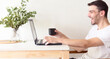 © Pablo - Retrato de un hombre adulto y apuesto caucásico sentado frente a su laptop y bebiendo una taza de café mientras la usa en un apartamento brillante y blanco con luz natural.