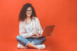 © denis_vermenko - Happy young curly beautiful woman sitting on the floor with crossed legs and using laptop on red background.