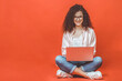 © denis_vermenko - Happy young curly beautiful woman sitting on the floor with crossed legs and using laptop on red background.