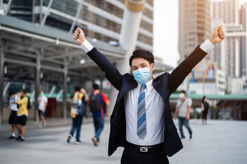 Wall Mural - successful business man with arms raised and wearing medical mask during coronavirus (covid-19) pandemic in city