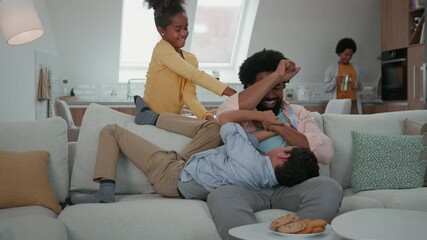 Wall Mural - Afro family in the living room. Father tickling his children, while mother is behind them in kitchen