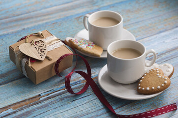 Valentine's day greeting card: two cups of coffee with milk on a blue wooden background, heart-shaped cookies on saucers, a gift box with a wooden heart, a red ribbon