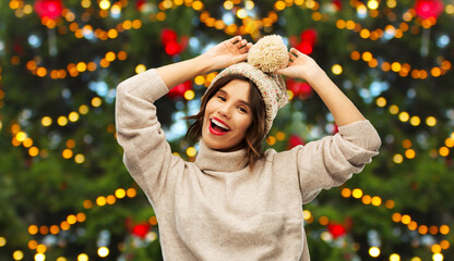 Sticker - winter holidays and people concept - happy smiling young woman in knitted hat and sweater over christmas tree lights on background