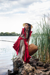 Wall Mural - beautiful young woman in red clothes and hat in asian style, with black hair and red braids, on the log by the river, fishing rod