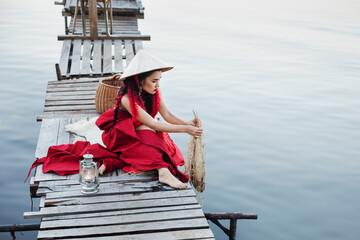 Wall Mural - beautiful young girl woman in designer clothes and hat in asian style, with black hair and red braids, is sitting on the pier and fishing