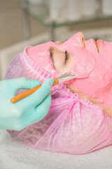 Wall Mural - Hand of cosmetologist applying pink alginic mask to face of young woman in beauty salon