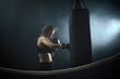 © producer - sporty brunette woman in boxing gloves and sportswear hitting the boxing bag in dark background with smoke.