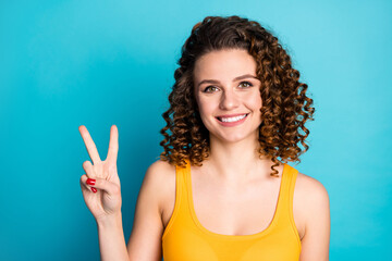 Poster - Photo of foxy pretty young girl raise hand show v-sign greeting beaming smiling wear yellow singlet isolated blue color background