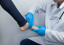 An Orthopedic Surgeon Examines A Woman's Leg. Foot Pain, Tendon Sprains, Inflammation, Flat Feet, Bursitis, Fasciitis. Foot Disease Treatment Concept. The Doctor Examines