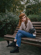 © sergeyzapotylok - Young thoughtful redhead woman in hoodie hand supports the chin enjoying warm fall day sitting in city park with green and yellow foliage background