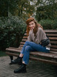 © sergeyzapotylok - Young thoughtful redhead woman in hoodie hand supports the chin enjoying warm fall day sitting in city park with green and yellow foliage background