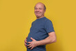 © Viktor Koldunov - Fat senior man touching his big abdomen with hands after well eating. Overeating problem. Studio shot on yellow wall.