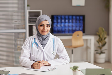 Wall Mural - Portrait of young female doctor in white coat looking at camera while sitting at the table at office