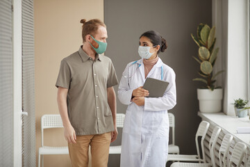 Wall Mural - Young nurse and patient in protective masks looking at each other while walking along the corridor at clinic