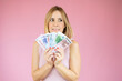 © Danko - Image of young funny woman standing over pink wall wearing casual clothes holding money. Looking aside.