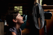 Young Boy With Eyes Closed Cooling Off In Front Of A Fan In Dark Room.