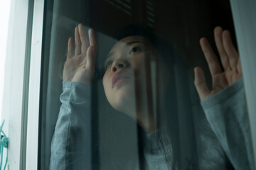 Wall Mural - young sad and depressed Asian Chinese girl scared and worried during covid19 lockdown and stay home quarantine looking through window feeling unhappy and stressed