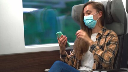 Wall Mural - Young woman passenger in a medical mask with phone while safety travel by train. Virus protection and wearing mask in public transport