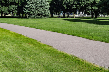 Asphalt footpath going straight throuhg a city park lawn with trees and a street in the background