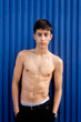 © Cavan Images - Young shirtless man hands on pocket, leaning on blue wall