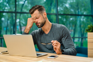 tired young businessman sitting at office desk with laptop with his eyes closed after computer work