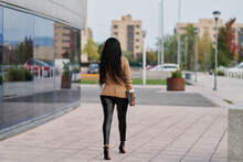 Back View Of A Black Woman Wearing A Brown Suit Holding A Laptop And A Container Of Coffee Walking Next To A Building. Business Concept