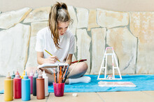 White Girl Painting A Picture Sitting On A Terrace With All Painting Elements Around As Brushes, Paintings, Water, Easel..