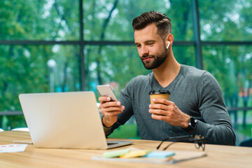 Smart modern young handsome businessman checking his phone during work break drinking coffee at office desk