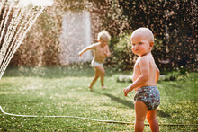Young Children Playing With Water From Sprinkler In The Backyard